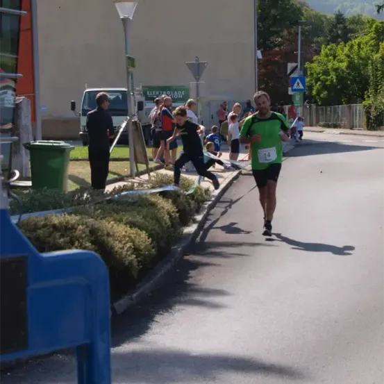 Ein Marathonläufer mit der Nummer 14 auf dem Trikot sprintet die Straße hinunter. Menschen versammeln sich am Straßenrand. Ein Lkw und eine Straßenlaterne sind in der Nähe.