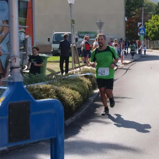 Ein Mann in einem grünen Shirt mit der Nummer 14 läuft auf einer Straße mit einer blauen Barriere und Büschen, in der Nähe eines Gebäudes mit einem Lastwagen und Menschen.