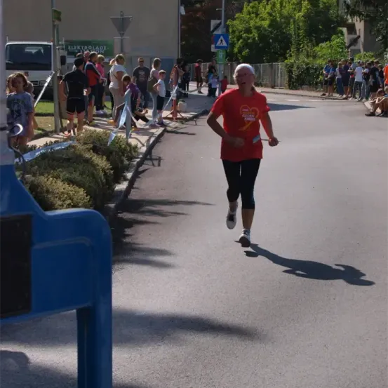 Ein Läufer in einem roten Shirt läuft auf einer Straße, umgeben von Zuschauern und Grünflächen. Die Straße hat eine weiße Linie und eine blaue Barriere. In der Ferne befindet sich ein LKW und ein Gebäude.