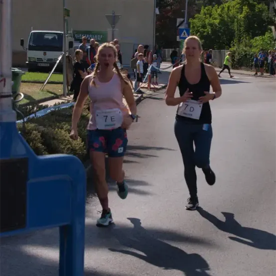 Zwei Frauen laufen auf der Straße während eines Marathons. Eine trägt ein rosa Shirt mit der Nummer 7E, die andere ein schwarzes Shirt mit der Nummer 7D. Hinter ihnen stehen Bäume und ein geparkter LKW.