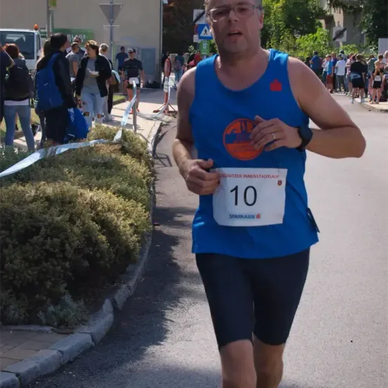 Ein Mann in einem blauen Tanktop und schwarzen Shorts läuft einen Marathon mit der Nummer 10 auf seinem Shirt. Zuschauer stehen hinter einer Seilbarriere im Hintergrund.
