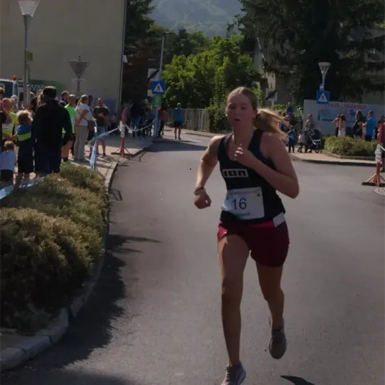 Eine Läuferin mit der Nummer 16 nimmt an einem Marathon teil, der auf einer Straße mit Büschen und Zuschauern in der Nähe läuft.