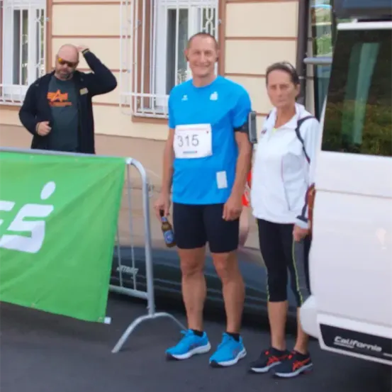 Ein Mann mit einer Flasche, ein Läufer und eine Frau stehen vor einem Gebäude. Der Läufer trägt ein blaues Shirt mit der Nummer 315. Hinter ihnen ist ein grüner Banner.