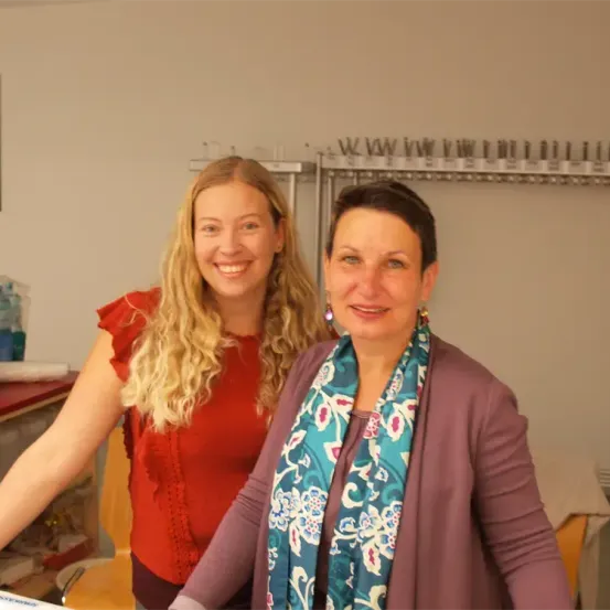 Zwei Frauen stehen lächelnd in einem Raum, eine trägt ein rotes Oberteil und die andere ein lilafarbenes Cardigan mit einem Blumenschal. Hinter ihnen ist eine weiße Wand mit einem Metallregal und einem Bild.