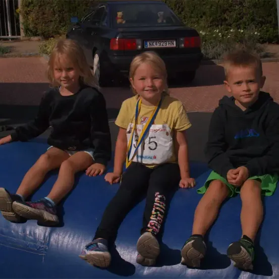 Drei Kinder sitzen auf einer blauen aufblasbaren Oberfläche. Ein Mädchen in der Mitte trägt ein gelbes Shirt mit der Nummer 375. Dahinter ist ein schwarzer Wagen auf der Straße geparkt.