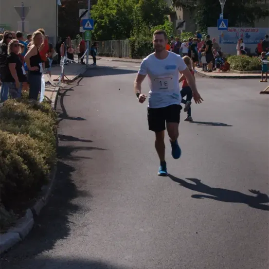 Ein Mann mit einem weißen Shirt und der Nummer 1E läuft einen Marathon. Zuschauer beobachten von der Seite. Bäume und Straßenschilder sind im Hintergrund.