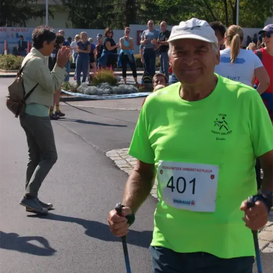 Ein älterer Mann in einem grünen T-Shirt mit der Nummer 401 steht auf der Straße, lächelt und hält einen Stock. Hinter ihm geht eine Frau, und mehrere Leute stehen und beobachten im Hintergrund.