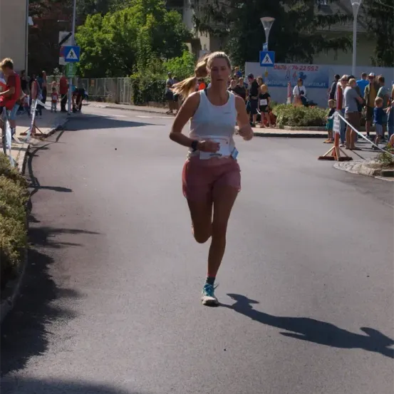 Eine Frau läuft an einem sonnigen Tag bei einem Marathon und trägt ein weißes Tanktop und pinke Shorts. Zuschauer stehen am Straßenrand.