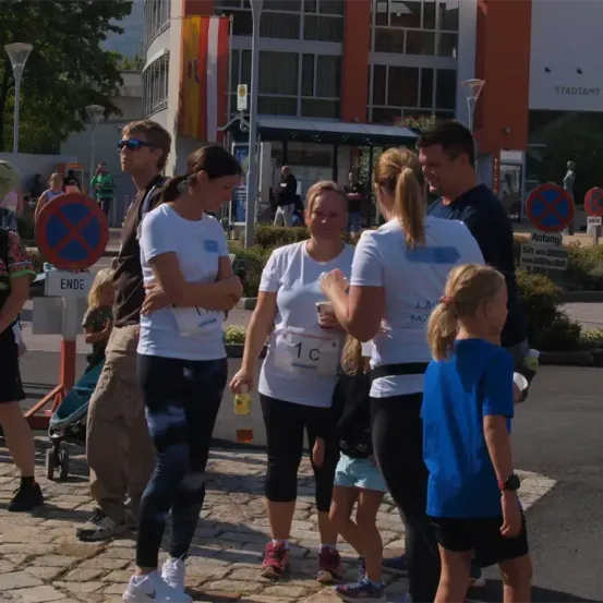 Vier Erwachsene und zwei Kinder versammeln sich in der Nähe eines Gebäudes mit einer Flagge, umgeben von Bäumen, Büschen und Schildern. Sie sind in weißen T-Shirts und Turnschuhen gekleidet, einige tragen Sonnenbrillen.