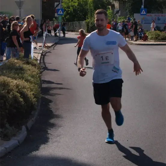 Ein Mann läuft bei einem Marathon und trägt ein weißes T-Shirt mit der Nummer 1E. Zuschauer stehen am Straßenrand und beobachten ihn.