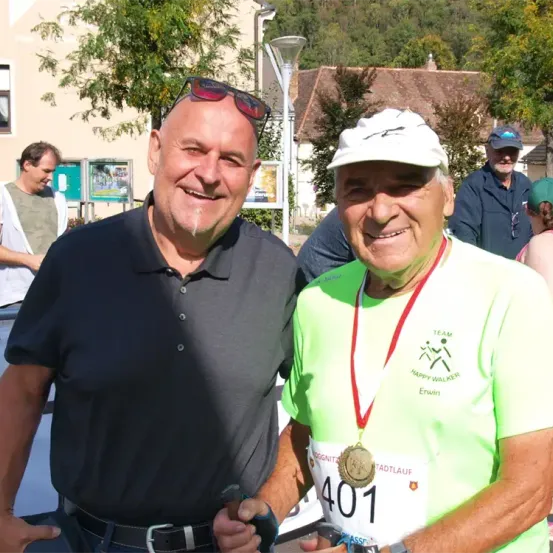 Zwei Männer stehen zusammen mit einer Medaille, einer von ihnen lächelt und trägt ein graues Shirt, der andere trägt ein grünes Shirt mit einer Medaille um den Hals.