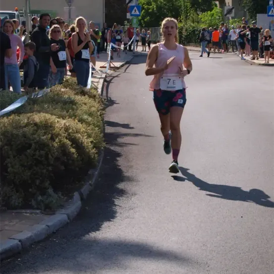 Eine Läuferin mit der Nummer 7E läuft auf einer Straße. Hinter ihr steht eine Menge Menschen und schaut zu. Einige tragen eine Brille, und einige halten Handys in der Hand.