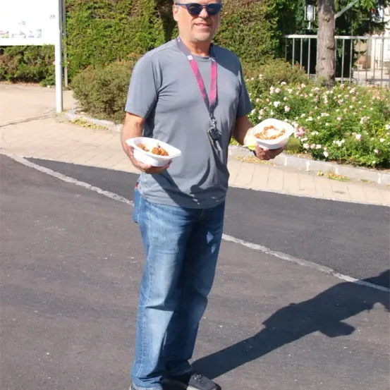 Ein Mann mit Sonnenbrille und grauem T-Shirt hält zwei Essensbehälter in einem Park. Er steht auf einer Straße mit Gehweg. Hinter ihm steht ein Schild und rechts davon ein Metallzaun.