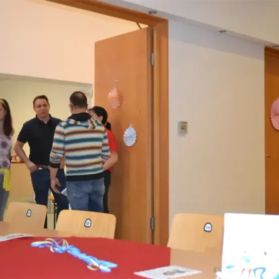 Mehrere Personen stehen in einem Raum mit einem rot bedecken Tisch. Einige schauen sich gegenseitig an, während andere nahe einer Wand mit Dekorationen stehen.