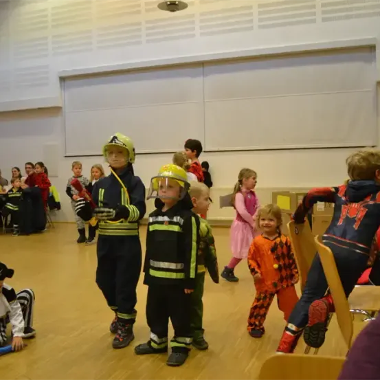 Mehrere Kinder sind in verschiedenen Kostümen gekleidet, darunter Feuerwehruniformen, in einem Raum mit Holzböden. Einige Kinder stehen, während andere auf Stühlen sitzen. Eine Frau hält in der Ferne ein Kind.