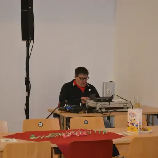 Ein Mann in einer schwarzen Jacke sitzt an einem Tisch, möglicherweise ein DJ, mit diversen Audiogeräten. Der Tisch hat eine rote Tischdecke, ein Buch und einen Becher. Ein Lautsprecher ist an der Wand montiert.