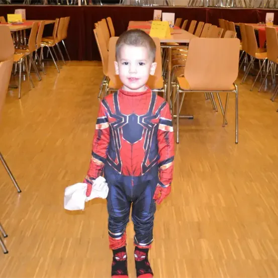 Ein kleiner Junge in einem Spiderman-Kostüm steht in einem Speisesaal mit Tischen und Stühlen. Er hält ein weißes Tuch in der linken Hand.