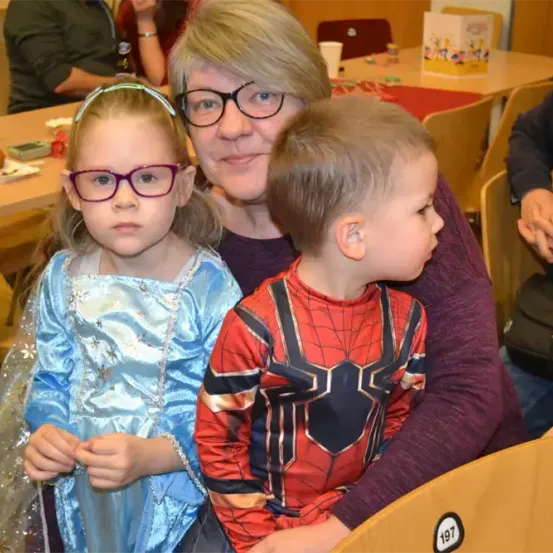 Eine Frau und zwei Kinder sitzen zusammen an einem Tisch, die Frau trägt eine Brille und die Kinder sind in Kostümen gekleidet. Ein Mädchen in einem blauen Kleid und ein Junge in einem Spiderman-Kostüm.
