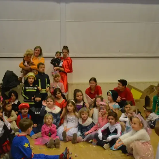 Eine Gruppe von Kindern in verschiedenen Kostümen ist in einem Raum versammelt, einige sitzen auf dem Boden, andere stehen. Ein Kind hält eine Puppe und es gibt Erwachsene im Hintergrund, einer hält ein Baby.