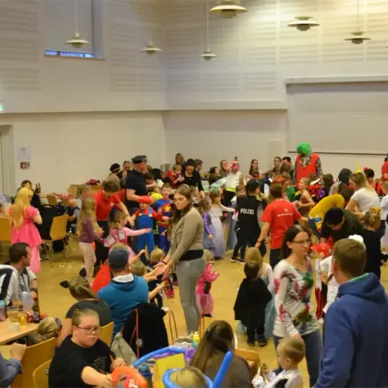 Eine große Gruppe von Menschen, darunter Kinder und Erwachsene, ist in einem Raum mit festlicher Atmosphäre versammelt. Viele sind in Kostümen gekleidet, einige mit Masken, andere in alltäglicher Kleidung. Der Raum ist mit Tischen und Stühlen gefüllt, und mehrere Personen stehen in der Nähe eines Tisches mit Luftballons.