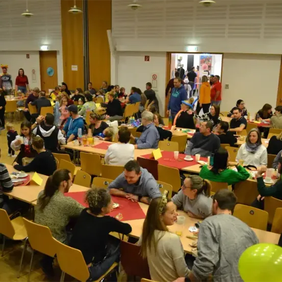 Eine große Gruppe von Menschen ist in einem Raum versammelt, um Tische mit Essen und Getränken herum sitzend. Viele tragen Hüte und Kostüme, und der Raum ist mit Luftballons und Schildern dekoriert.