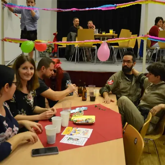 Eine Gruppe von Menschen sitzt um einen Tisch mit Ballons und Luftschlangen in einem Festsaal. Sie lächeln und scheinen die Veranstaltung zu genießen.