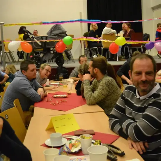 Eine Gruppe von Menschen sitzt an Tischen in einem Raum mit Luftballons und Girlanden. Einige lachen und reden, während andere sich umsehen. Ein Mann lächelt direkt in die Kamera.