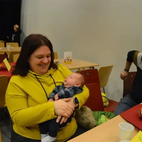Eine Frau mit einem lächelnden Gesicht hält ein Baby in den Händen. Eine Person, die neben ihr sitzt, schaut etwas an. Der Ort scheint ein Restaurant zu sein. Ein Tisch mit einer Tasse und einer Schachtel steht hinter ihnen. Eine andere Person sitzt auf dem Stuhl hinten.