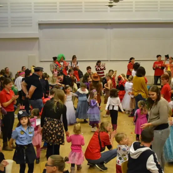 Eine Gruppe von Menschen, darunter Kinder in Kostümen, ist in einem Raum versammelt, wahrscheinlich für einen Karneval oder eine Party. Einige stehen, andere sitzen auf dem Boden. Eine Frau in einer roten Uniform und einem Hut steht neben einer Person in einem Clownskostüm. Einige Leute halten Luftballons.