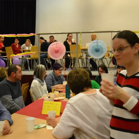 Eine Gruppe von Menschen sitzt an Tischen in einem Raum mit Luftballons und Girlanden. Eine Frau steht und hält einen Becher. Eine andere Frau sitzt an einem Tisch mit Essen und Getränken.