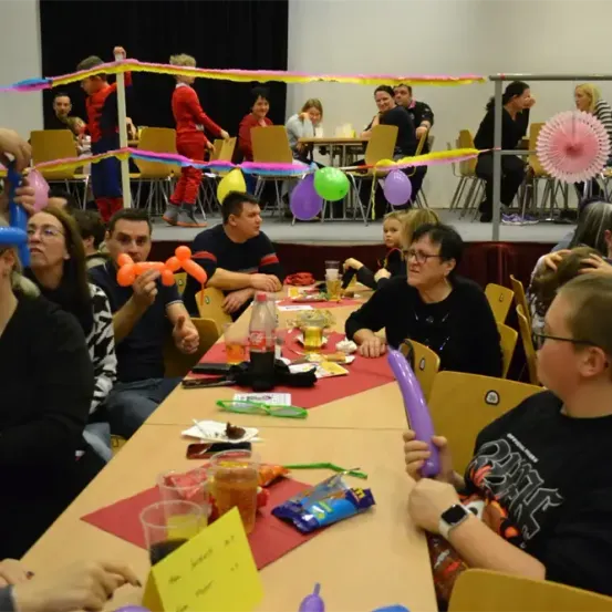 Eine große Gruppe von Menschen auf einer Party. Luftballons und Dekorationen schmücken den Bereich. Einige sitzen an Tischen mit Essen, Getränken und Spielen. Andere stehen und unterhalten sich. Kinder spielen auf der Bühne.
