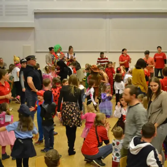Eine Gruppe von Menschen, einschließlich Kindern und Erwachsenen in Kostümen, versammelt sich in einem Raum, einige tanzen, während andere reden. Ein Mann in Uniform steht in der Mitte.