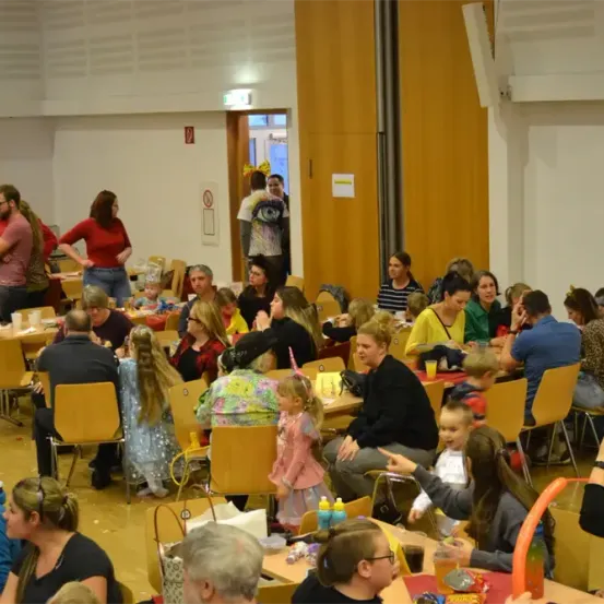 Eine große Gruppe von Menschen versammelt sich in einem Raum und sitzt an Tischen. Einige von ihnen sind Erwachsene, andere Kinder. Einige Kinder tragen Kostüme. Der Raum hat weiße Wände und Holzböden. Es gibt eine Tür und eine Lampe an der Decke.