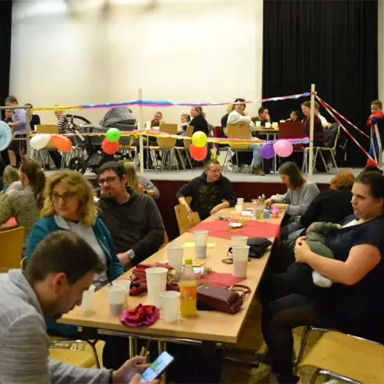 In einem Raum mit Tischen und Stühlen ist eine Gruppe von Menschen versammelt. Sie sitzen um Tische herum, die mit Bechern, Tellern und Luftballons besetzt sind. Ein Mann schaut auf sein Handy, und eine Frau hält ein Baby.