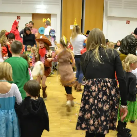 Eine lebhafte Szene im Innenbereich, bei der Kinder in verschiedenen Kostümen, darunter Hexen, Cowboys und andere, mit Erwachsenen tanzen. Der Raum hat Holzböden und eine Wand, die mit Dekorationen geschmückt ist.