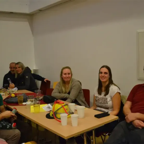 Eine Gruppe von Menschen versammelt sich um einen Tisch in einem Raum. Ein Baby sitzt auf dem Schoß eines Mannes in einem grünen Hemd. Eine Frau sitzt neben einem Feuerwehrhelm. Eine andere Frau hält ein Handy.