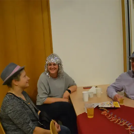 Drei Personen sitzen um einen Tisch herum und tragen Hüte. Eine Frau in der Mitte trägt eine silberne Paillettenperücke und lächelt. Zwei Gläser mit gelben Getränken stehen auf dem Tisch.