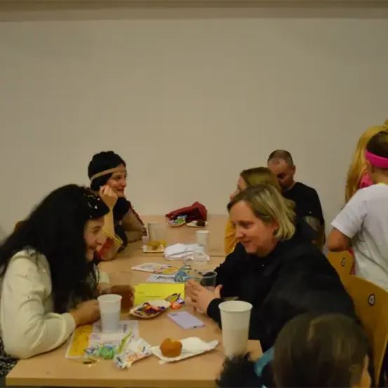 Eine Gruppe von Menschen, darunter Erwachsene und Kinder, sind um einen Tisch herum versammelt. Einige tragen Kostüme, während andere lässig gekleidet sind. Auf dem Tisch stehen Snacks, Getränke und verschiedene Gegenstände.