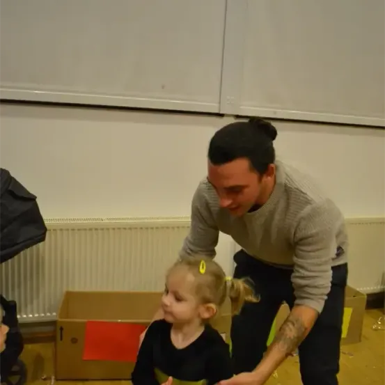 Ein Mann mit Tätowierungen beugt sich zu einem kleinen Mädchen hinunter, beide lächeln. Sie stehen in einem Raum mit weißer Wand, Heizkörper und Holzboden.