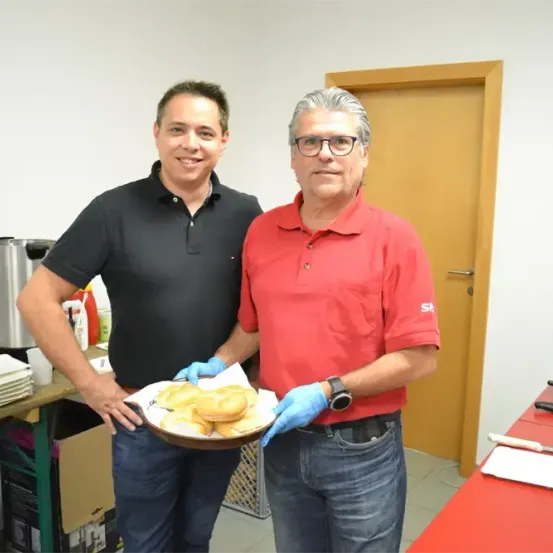 Zwei Männer stehen in einer Küche. Einer hält eine Schüssel mit Essen, beide tragen Handschuhe. Ein Tisch und eine Tür sind hinter ihnen.