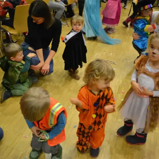 Mehrere Kinder in Kostümen stehen auf einem Holzboden. Ein Kind in einem grünen Kostüm schaut ein anderes an, das ein orangefarbenes trägt. Dahinter beobachtet eine Frau.