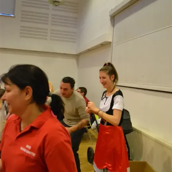 Eine Frau in einem rot-weißen Kleid tanzt mit einer anderen Frau in einem roten Shirt. Ein Mann in einem beigen Pullover sitzt auf einem Stuhl hinter ihnen. Sie befinden sich in einem Raum mit weißen Wänden und einer Decke mit einer Lampe.