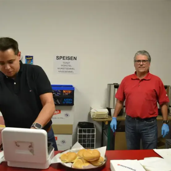 Zwei Männer in einer Cafeteria, einer serviert Essen, während der andere zusieht. Ein Schild zeigt 'Toast Wurstel Wurstsemmel Käsesemmel'.