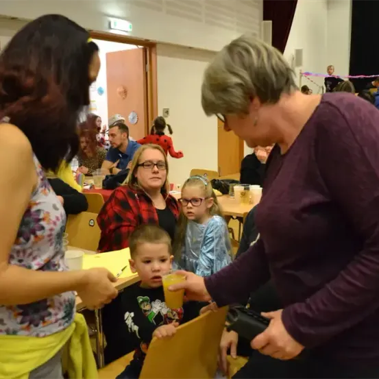 Eine Zusammenkunft in Innenräumen mit Erwachsenen und Kindern. Eine erwachsene Frau füttert einen jungen Jungen mit einem Getränk.