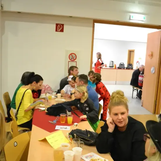 Eine Gruppe von Menschen sitzt um einen Tisch in einem Raum. Auf dem Tisch befinden sich verschiedene Gegenstände wie Getränke, Essen und ein Spiel. Der Raum hat weiße Wände und eine Tür auf der rechten Seite. In der Nähe der Tür steht eine Person.