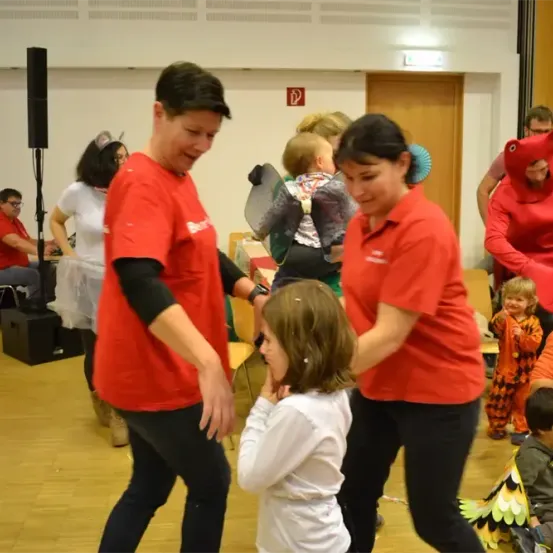Zwei Frauen in roten T-Shirts tanzen mit einem kleinen Mädchen in einem weißen T-Shirt. Dahinter schauen eine Gruppe von Menschen zu, darunter ein Mann in einem roten Dinosaurierkostüm.