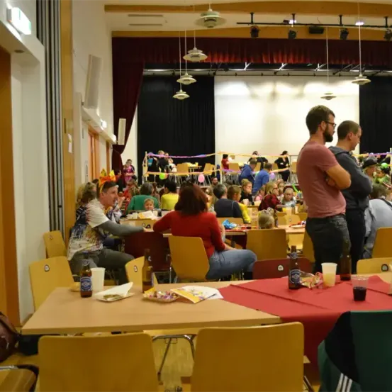 Ein Raum mit Tischen, die für ein Treffen vorbereitet sind. Menschen sitzen und stehen, einige unterhalten sich. Flaschen, Becher und Snacks sind auf den Tischen. Der Raum hat eine Bühne und Dekorationen.