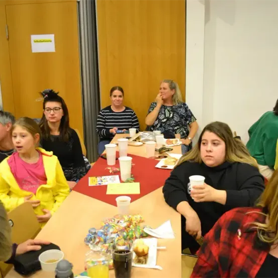 Eine Gruppe von Erwachsenen und Kindern sitzt in einem Raum um einen Tisch herum und isst. Einige halten Tassen und andere essen. Auf dem Tisch liegt ein Tischläufer und es gibt Teller, Tassen und andere Gegenstände darauf. Eine Frau schaut etwas an. Hinter ihnen befindet sich eine Tür mit einem Schild.