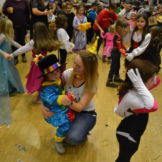 Mehrere Kinder in Kostümen tanzen auf einem Holzboden. Eine Frau hält ein Kind in einem Clown-Kostüm. Andere sind in Feen-, Superhelden- und anderen Kostümen.