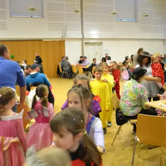 Eine Gruppe von Kindern in verschiedenen Kostümen versammelt sich in einem Raum mit Erwachsenen um sie herum. Die Kinder schauen auf etwas vor ihnen. Einige Erwachsene sitzen auf Stühlen, andere stehen. Es gibt Tische mit Essen darauf.